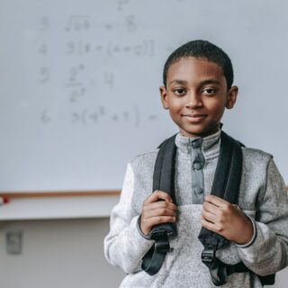 smiling black schoolboy near white board in classroom